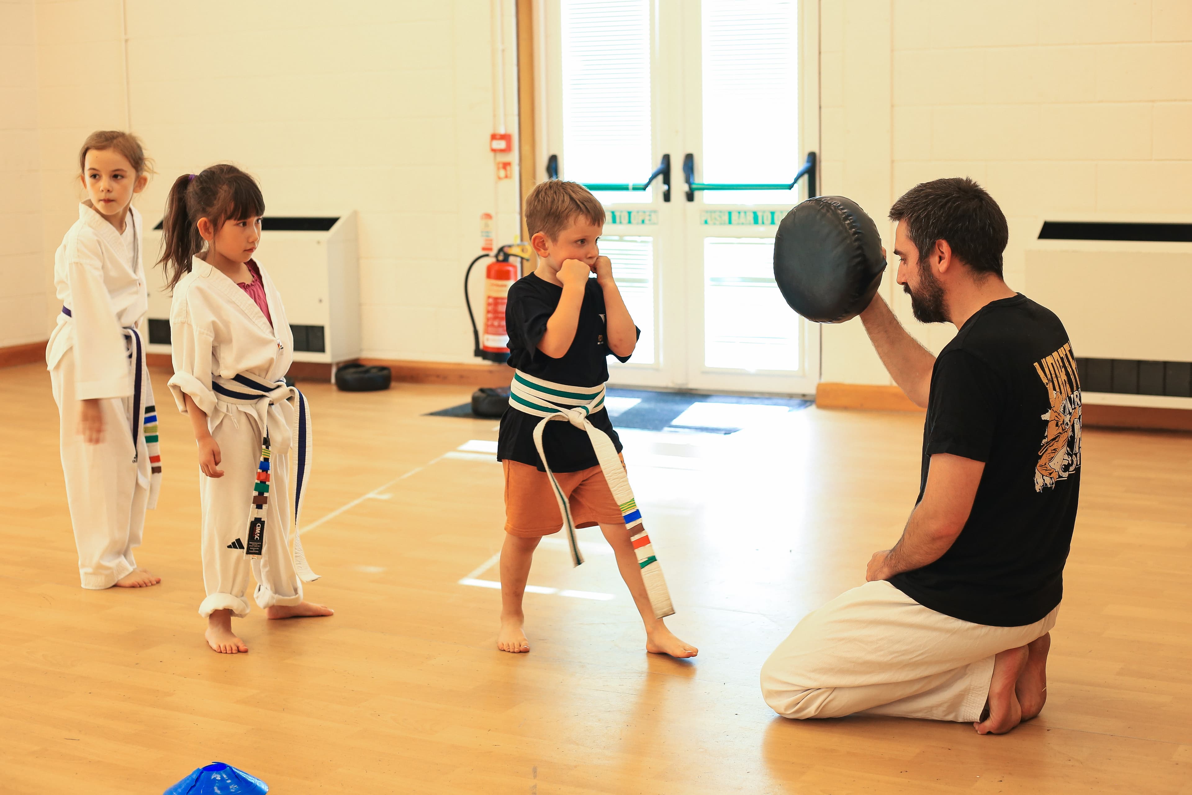 Dragons class at Vortex Martial Arts Academy, coach kneeling at kids' level showing kicking technique